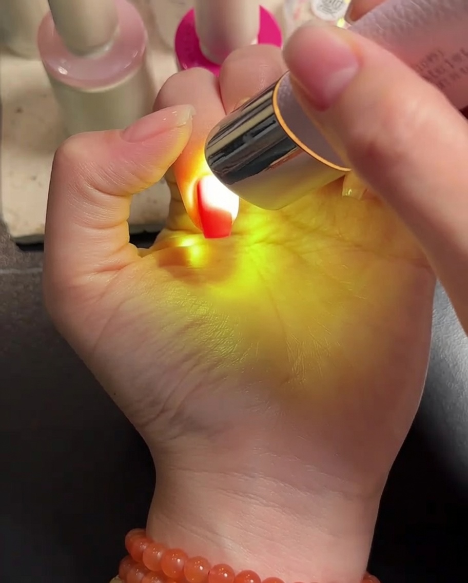 Une main placée sous une lampe LED portable, les ongles recouverts de gel étant catalysés.