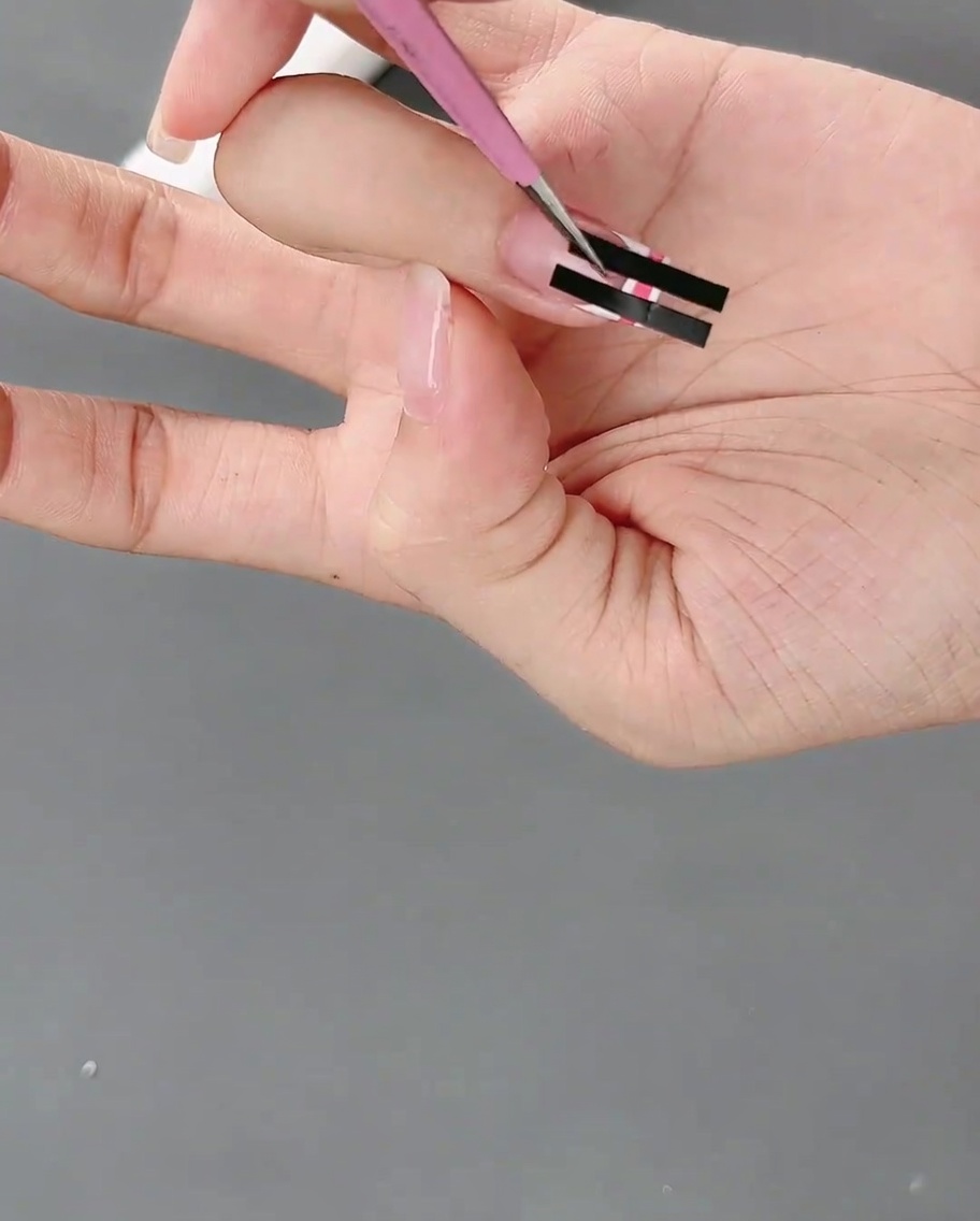 Une pince retire doucement un ruban adhésif dun ongle pour révéler un motif de grille à carreaux rose et blanc.