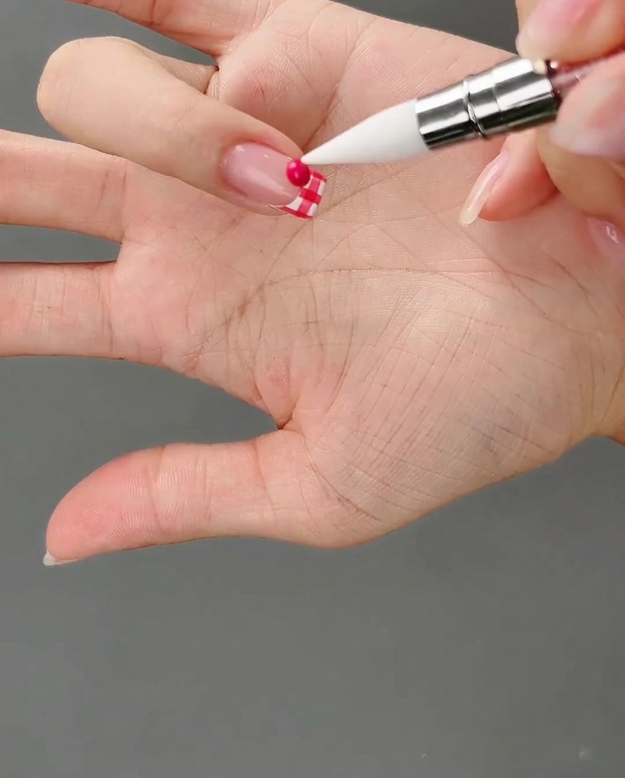 Un outil en silicone pointant vers une petite bille ronde de gel rose foncé posée sur le bout de longle quadrillé.