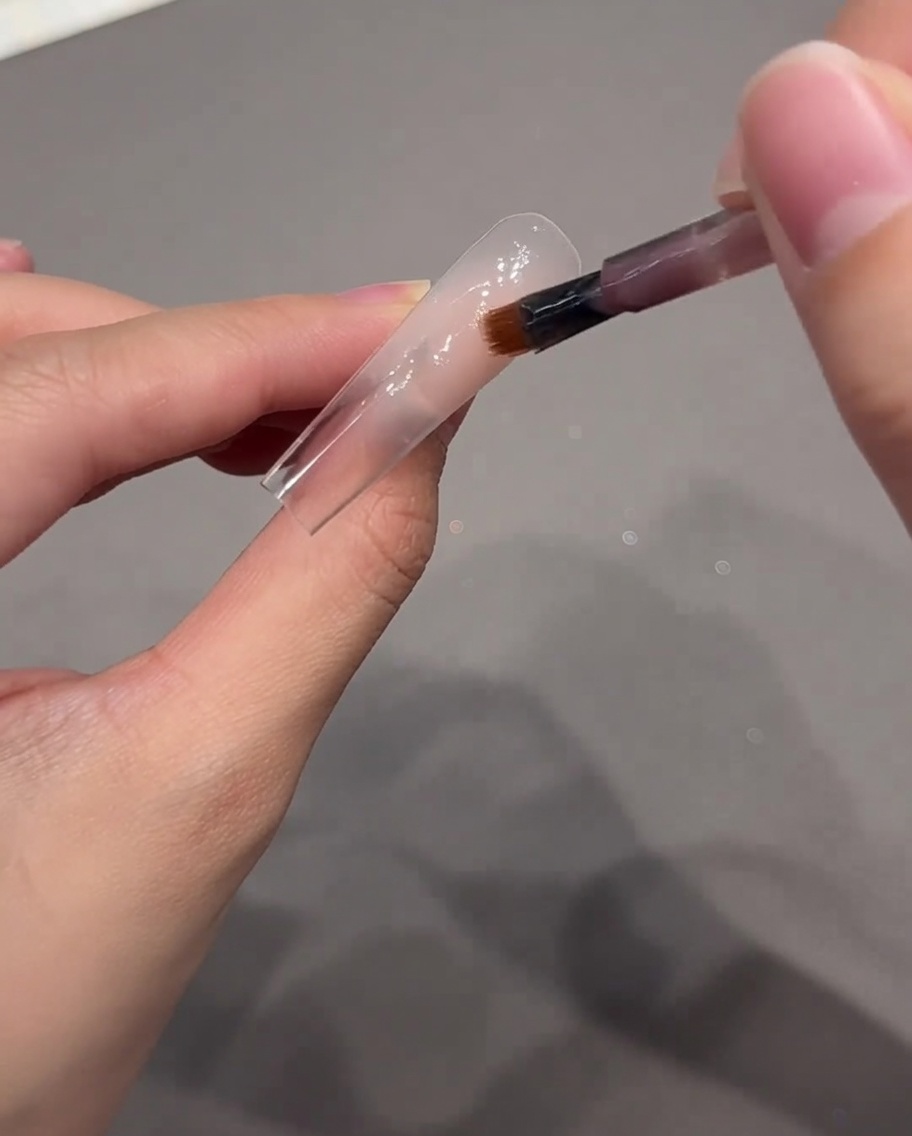 Un pinceau de nail art déposant une goutte de gel rose nude près de la cuticule dune capsule.