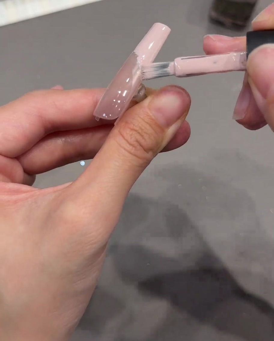 Un pinceau appliquant un gel rose nude opaque sur le bout dun ongle, utilisant un autocollant incurvé comme délimitation.