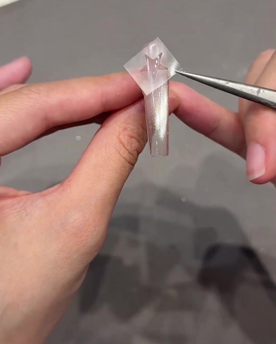 Une pince à épiler tenant un autocollant en forme détoile juste au-dessus dun ongle transparent de style cat-eye.