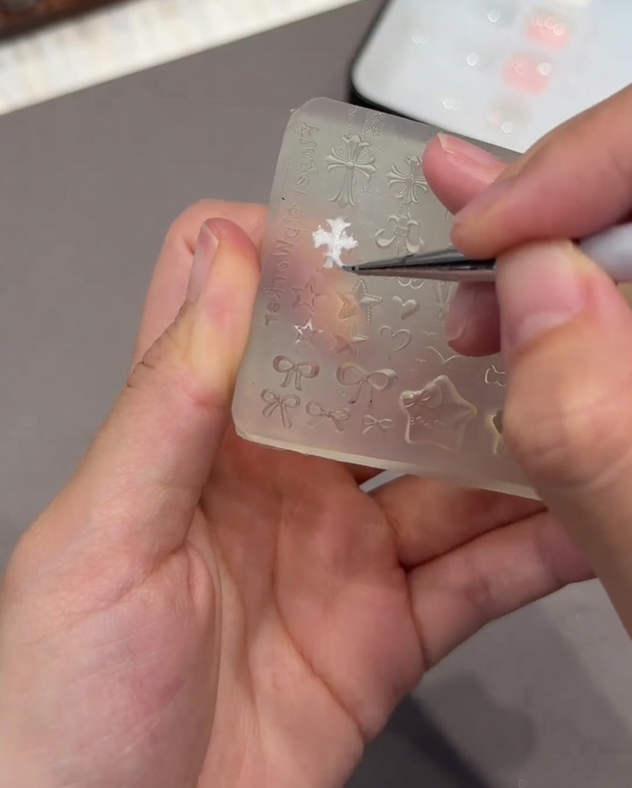 Une pince à épiler tenant un petit moule 3D blanc en forme de cœur, prête à le placer sur un ongle.