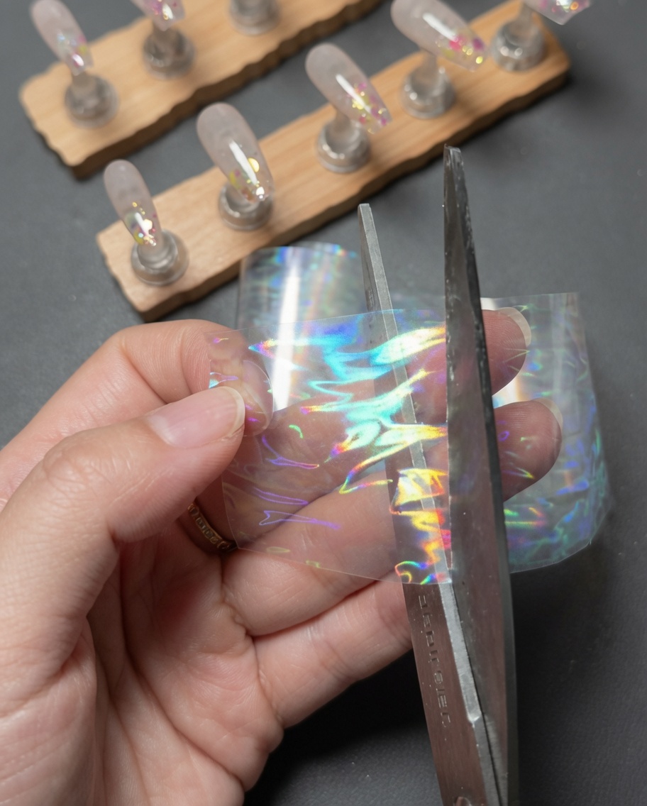 Des mains utilisant des ciseaux pour couper un petit morceau rectangulaire dans une bande de foil de transfert holographique aurore laser.