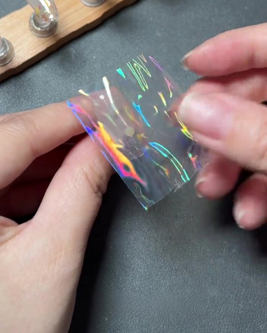 Des doigts plaçant soigneusement un petit morceau de foil de transfert holographique au centre dun ongle.