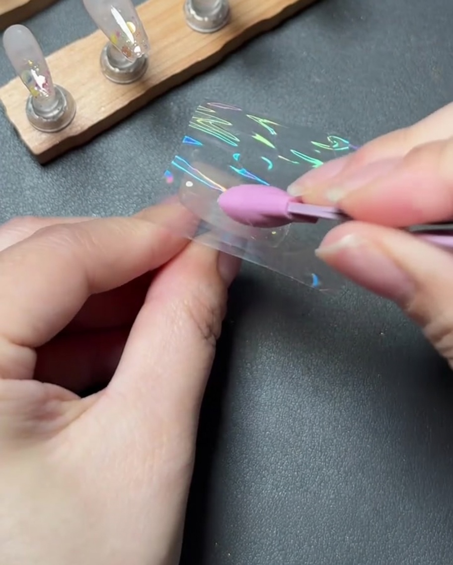 Un outil en silicone rose pressant et lissant fermement un morceau de foil de transfert holographique sur un ongle.