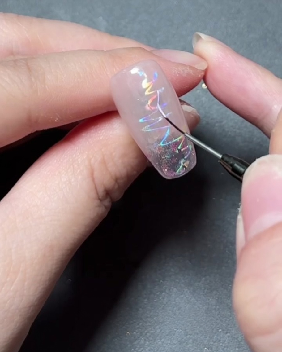 Un pinceau fin peignant une forme détoile délicate avec du gel coloré sur un ongle holographique.