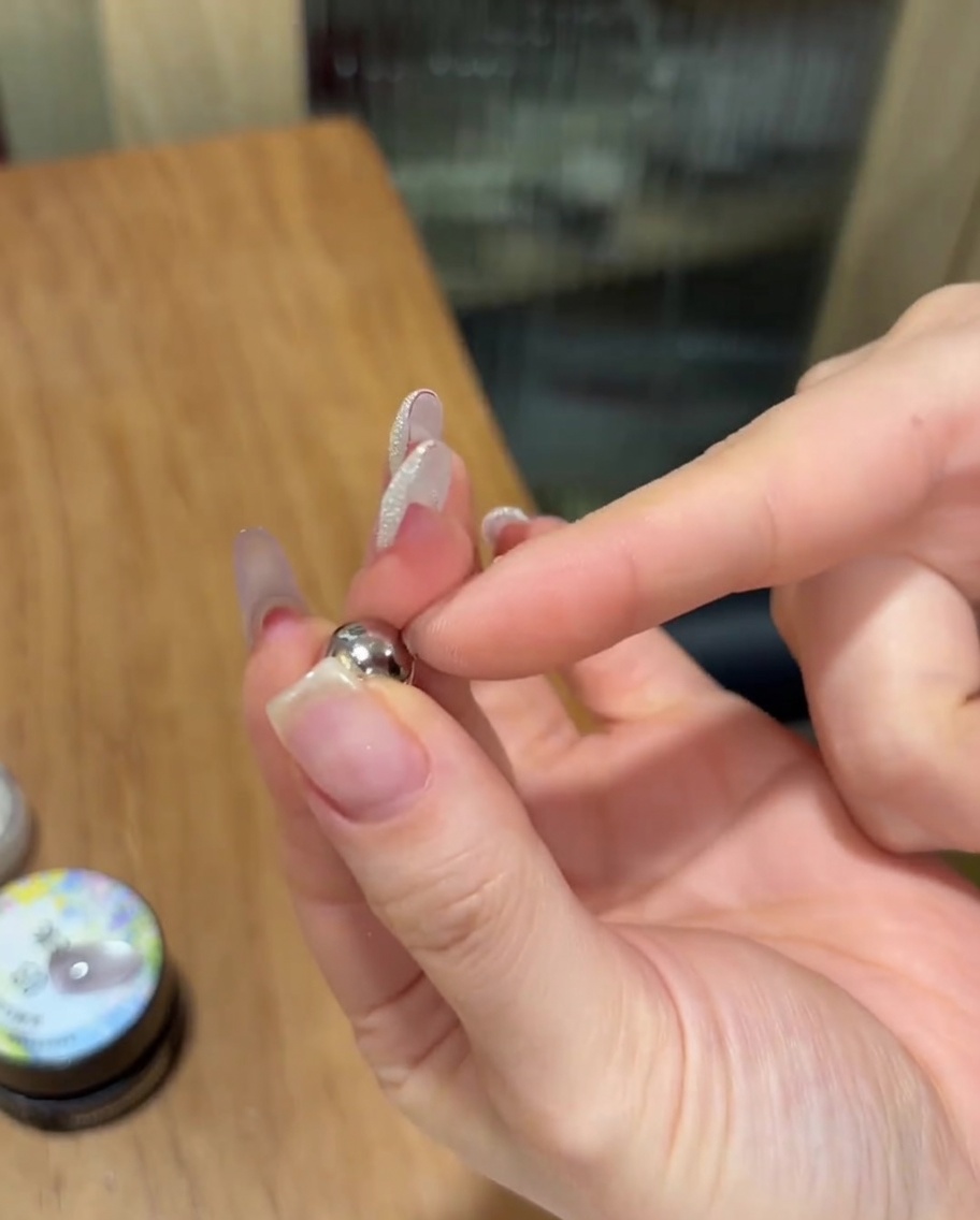 Une main tenant un aimant sphérique argenté près dun ongle avec du gel cat eye non catalysé.