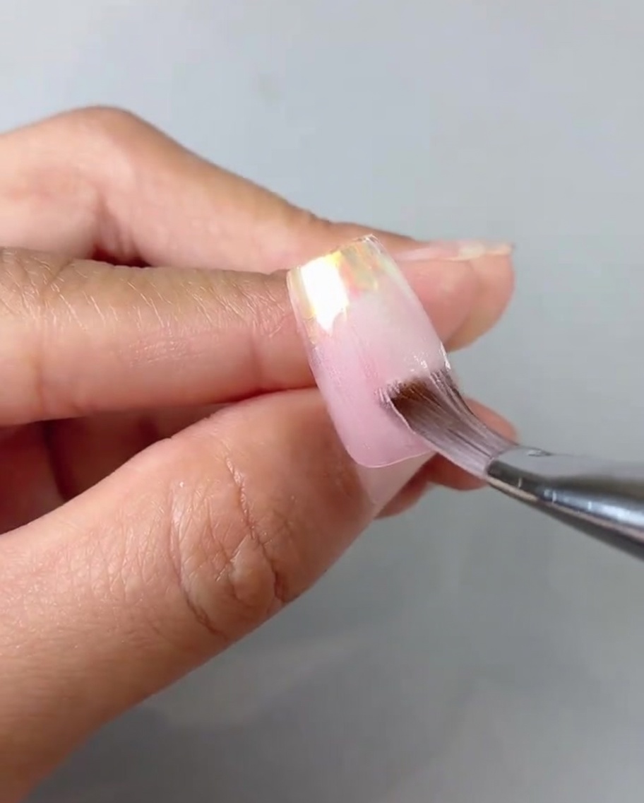 Gros plan dun pinceau lissant et estompant un gel rose translucide sur une capsule.