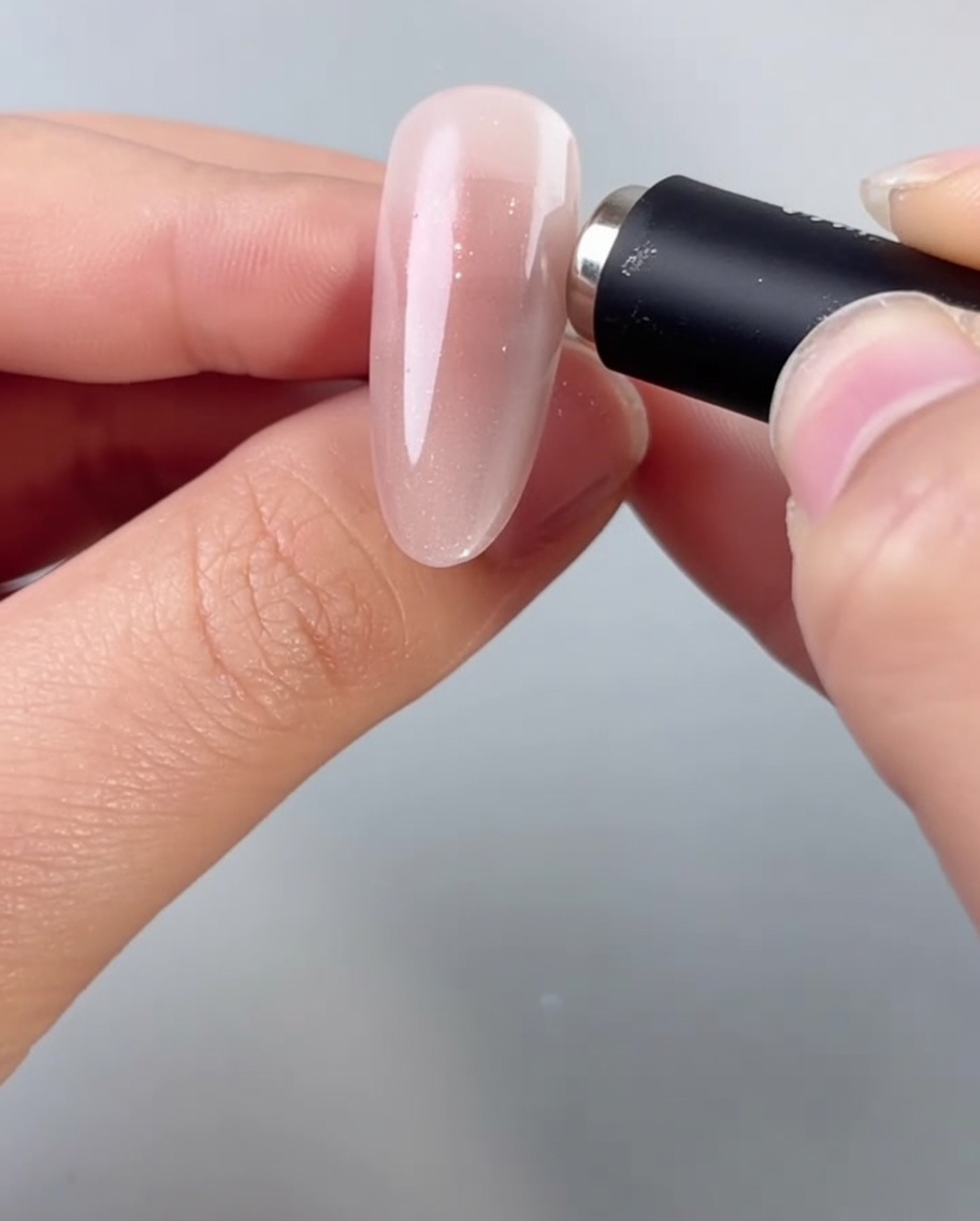 Une main tenant un aimant cylindrique près dun ongle en amande, activant les particules scintillantes du gel cat-eye humide.