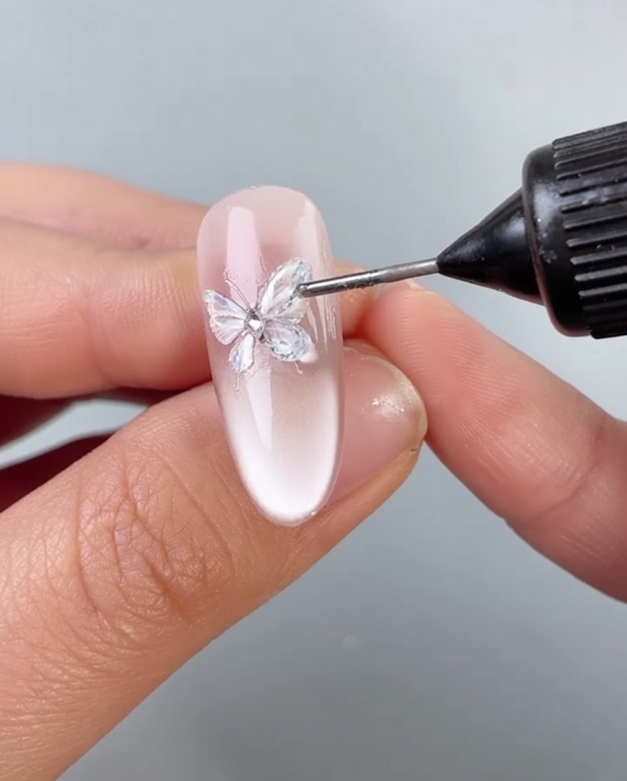 Un pinceau appliquant une fine couche de gel transparent autour de la base dune décoration papillon sur un ongle.