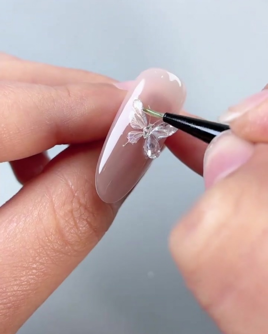 Gros plan dun pinceau appliquant un gel sculptant épais et transparent sur les ailes gauches dun sticker papillon décoratif sur un ongle rose.