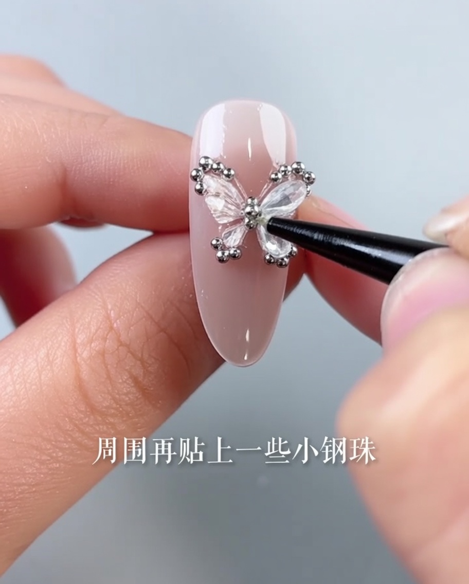 Une pince de précision plaçant de minuscules billes en acier argenté autour du bord dun ongle décoré dun papillon.