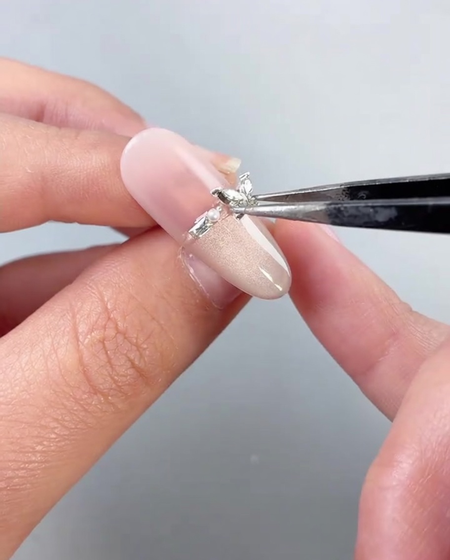 Une pince plaçant un bijou papillon métallique sur un ongle décoré.