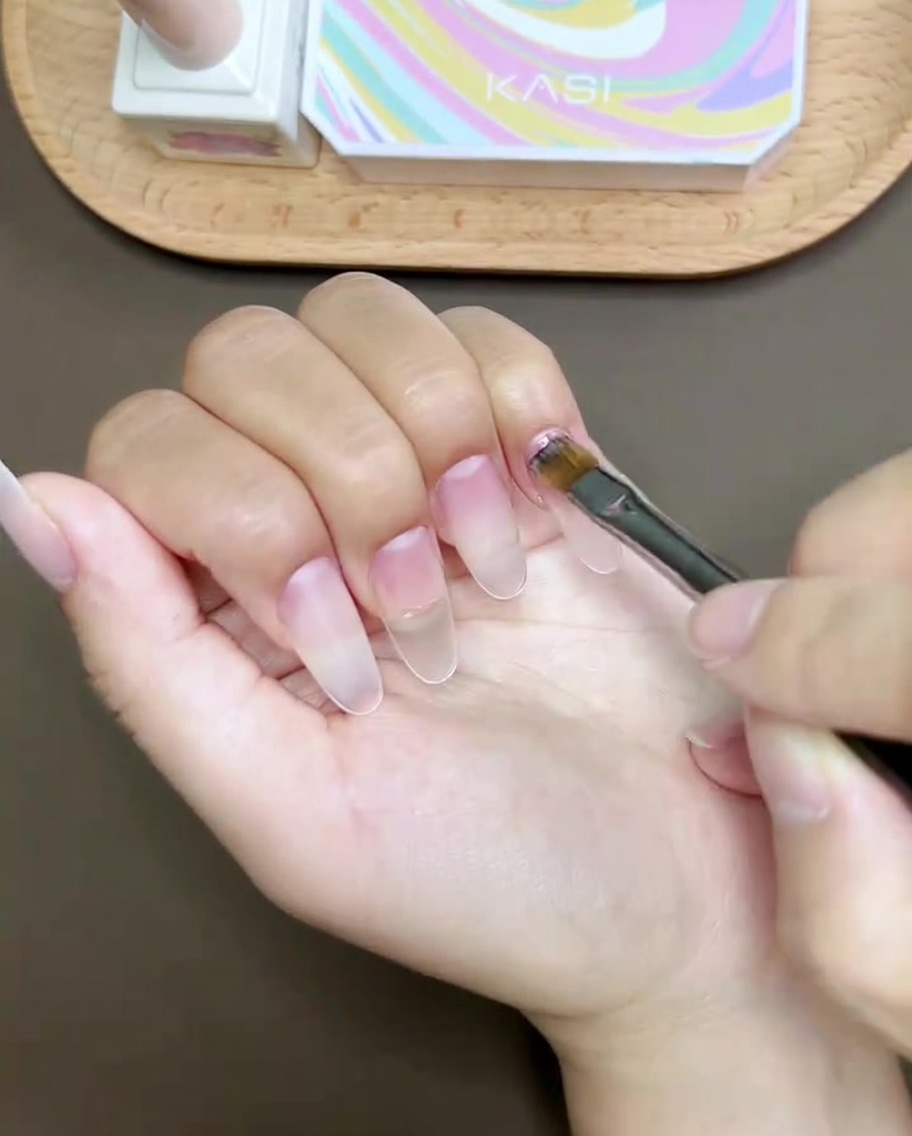 Un pinceau à ongles peignant une ligne courbe rose sur le bout de longle du pouce.