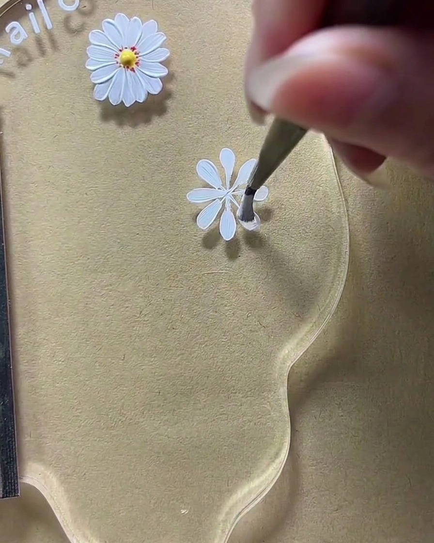 Gros plan sur une palette de nail art où un pinceau élargit les formes des pétales blancs sur un design de marguerite.