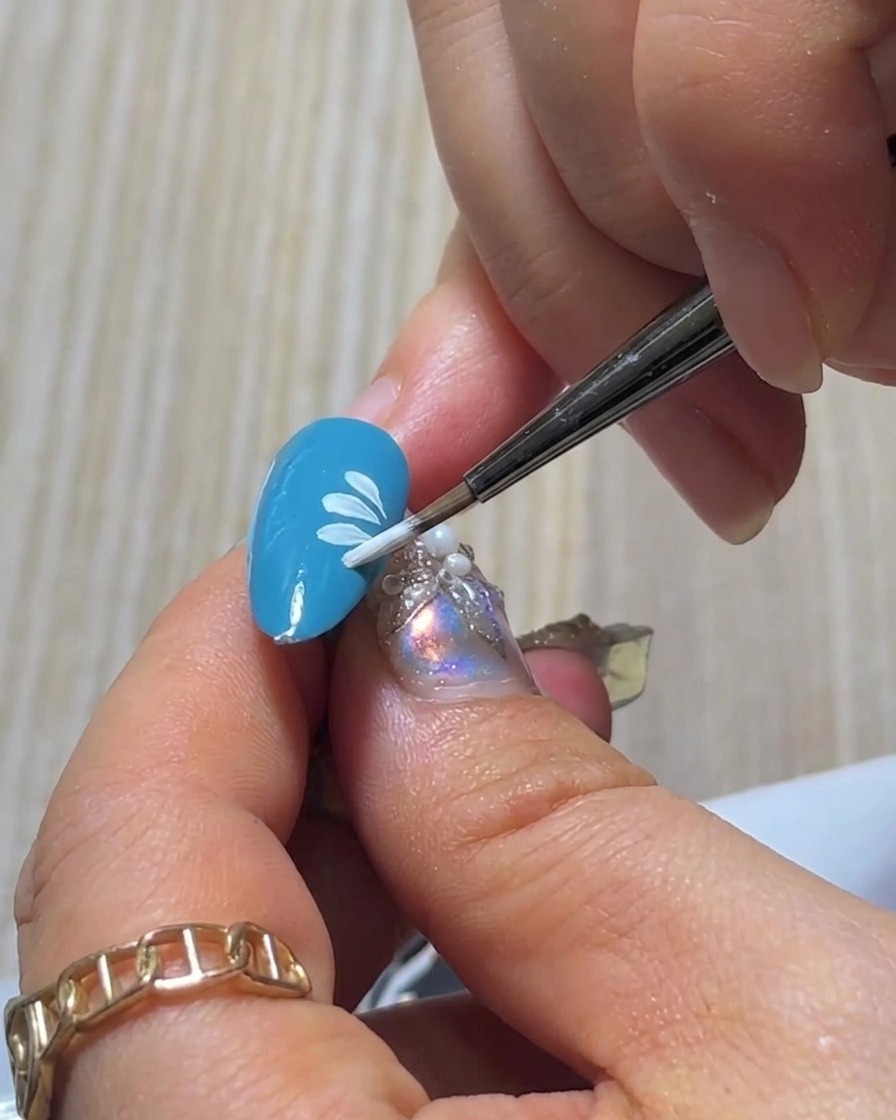 Une vue de près dun pinceau peignant le dernier pétale blanc pour compléter la couche inférieure dun petit motif de marguerite sur un ongle bleu.