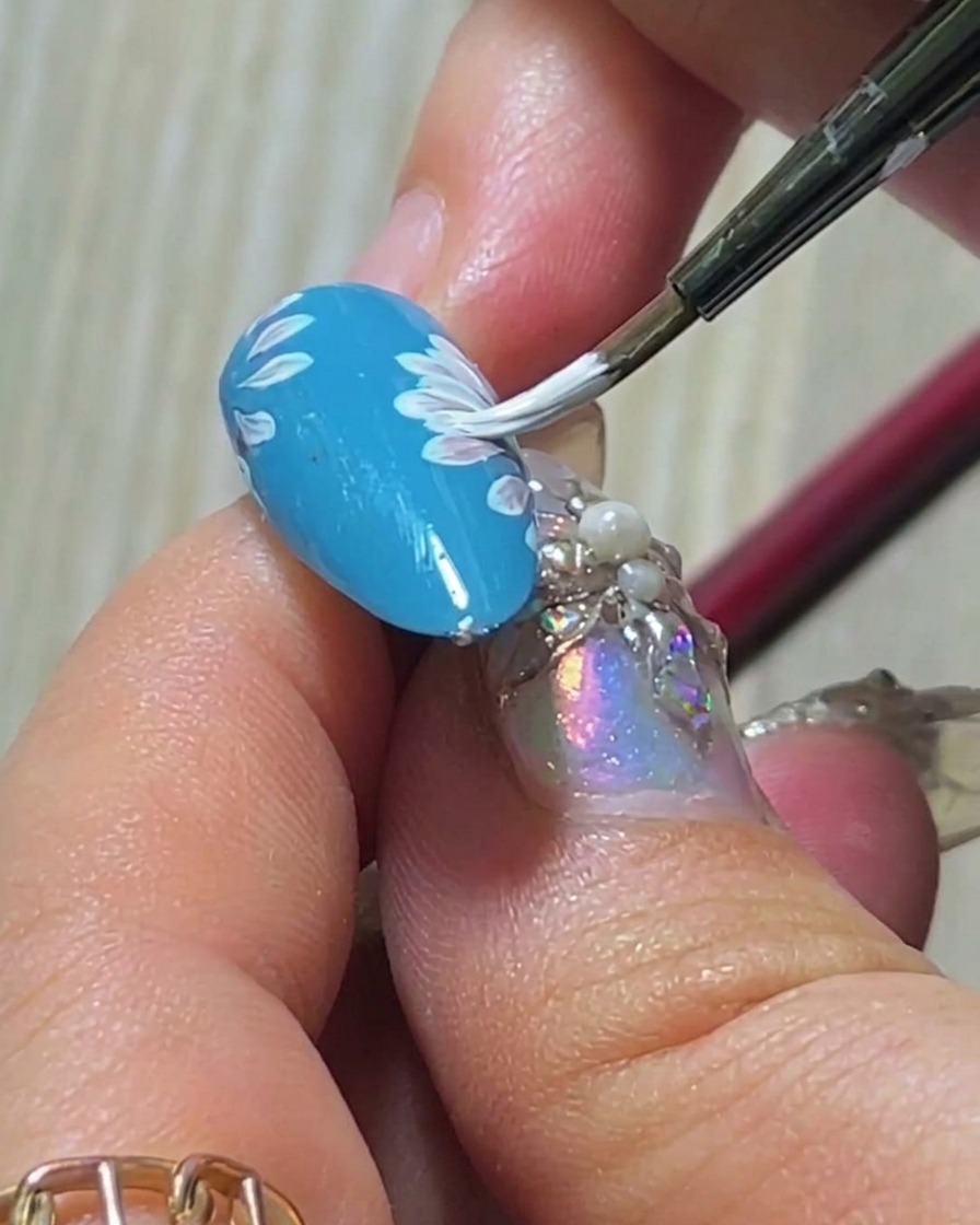 Un gros plan dun pinceau ajoutant un troisième pétale blanc à la couche superposée de la marguerite sur un ongle bleu.