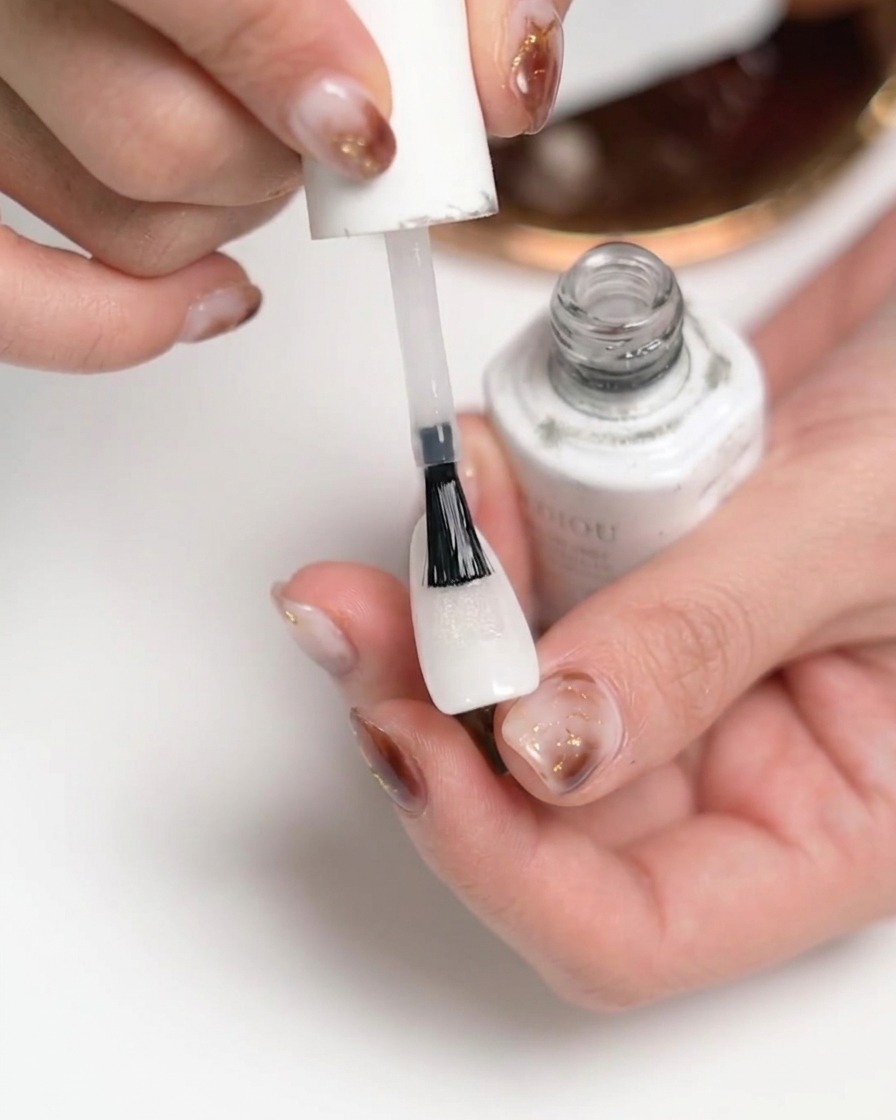 Gros plan dune main appliquant un top coat mat transparent sur un ongle peint en blanc.