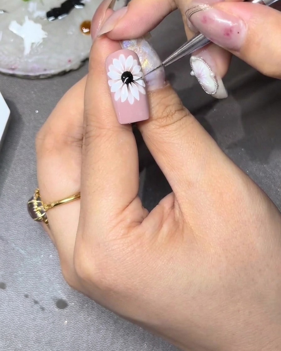 Gros plan dun ongle avec un motif floral blanc, où un pinceau fin étire de petits traits jaunes vers lextérieur depuis le centre dans les espaces entre les pétales blancs.