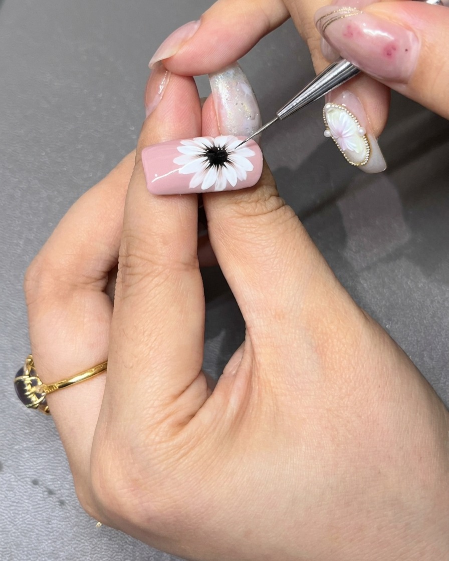 Un pinceau de détail fin affinant le centre noir foncé dune marguerite blanche peinte sur un ongle rose clair.