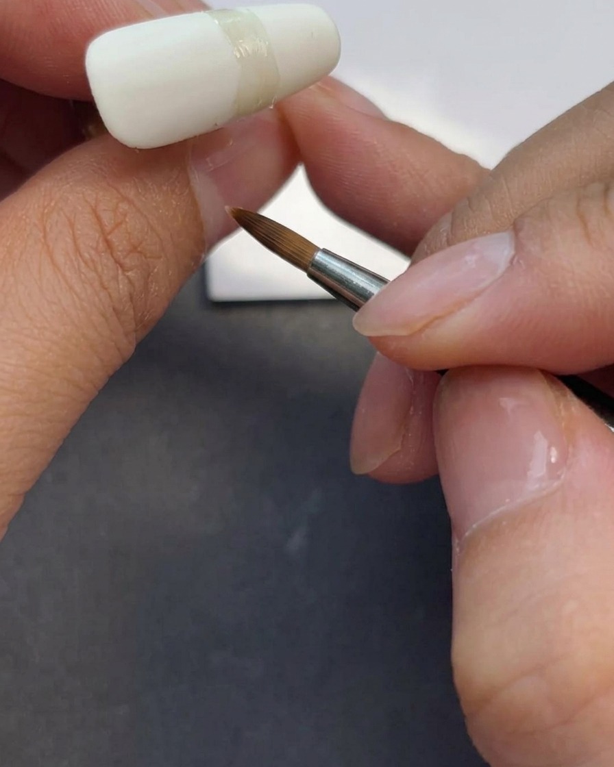 Une prothésiste ongulaire appliquant une couche de top coat mat sur un ongle blanc laiteux.