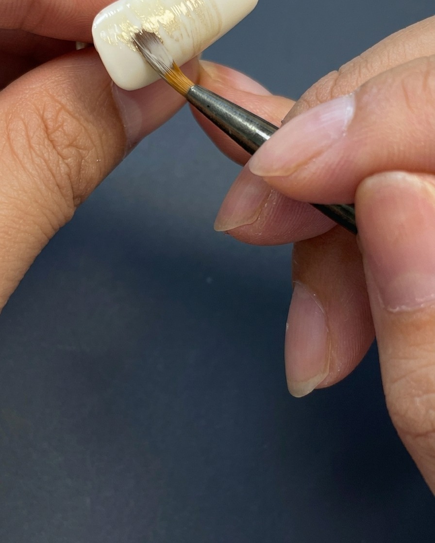 Un pinceau créant des traits horizontaux streaky avec un mélange de gel aux tons terreux sur un ongle blanc.