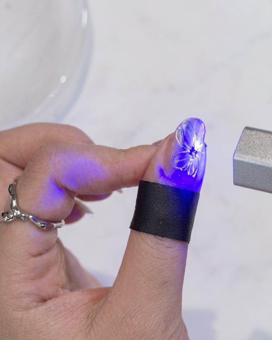 Un pinceau appliquant une dernière couche de top coat transparent sur un ongle présentant un design de fleur blanche avec un centre doré.