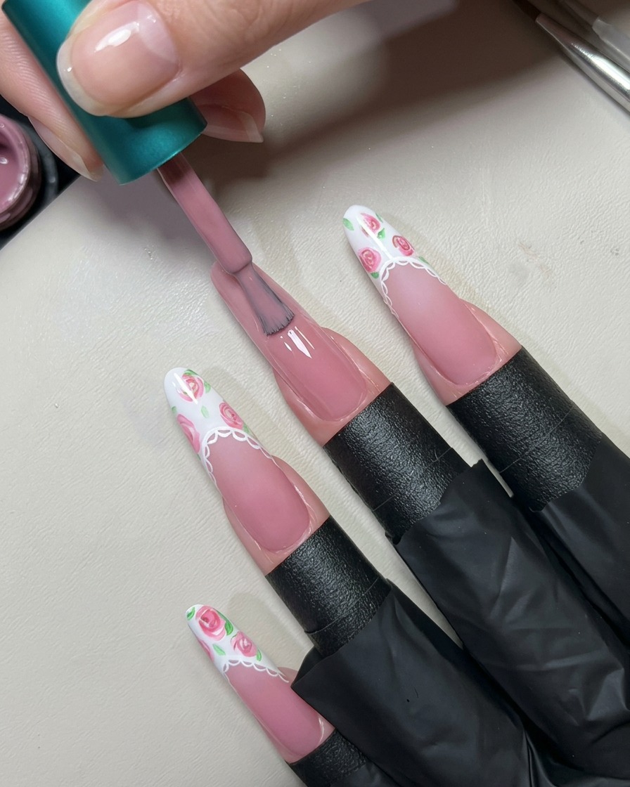 Un pinceau de vernis appliquant une couche de gel rose nude translucide sur un ongle long.
