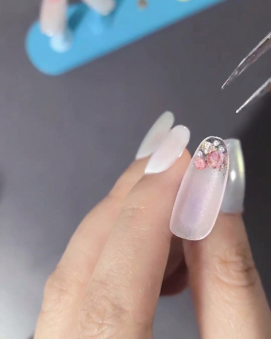 Gros plan dun ongle décoré de fleurs séchées, perles et petits strass métalliques.