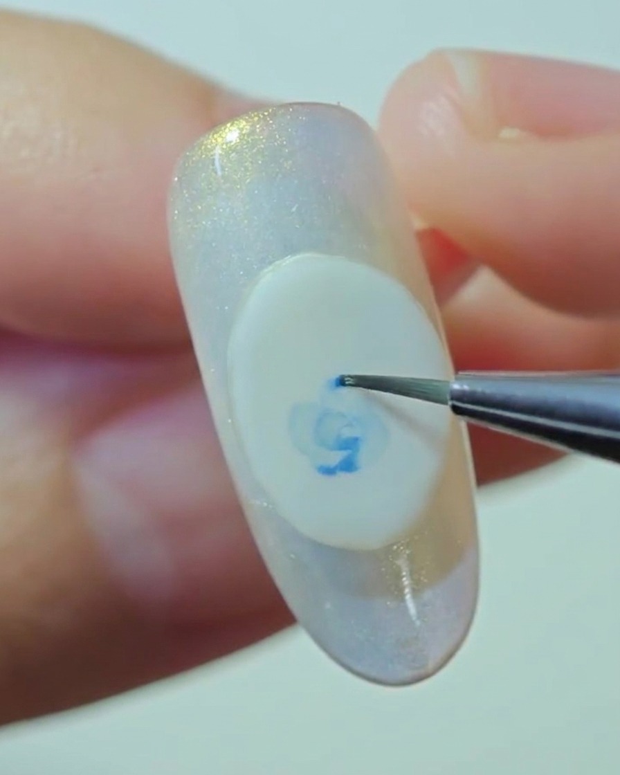 Gros plan sur un pinceau de détail mélangeant doucement les bords dune rose bleue sur une base en gel blanc.