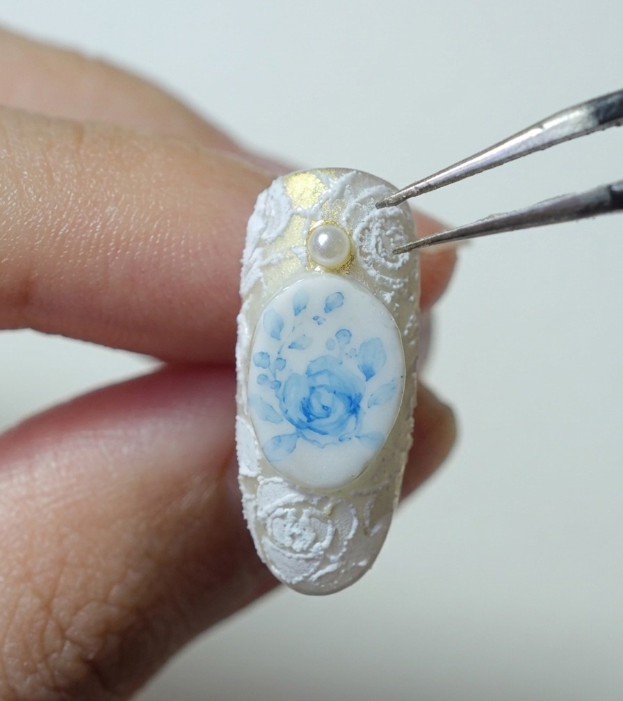 Une pince tenant une petite perle blanche au-dessus dun ongle avec art de rose bleue et dentelle blanche.