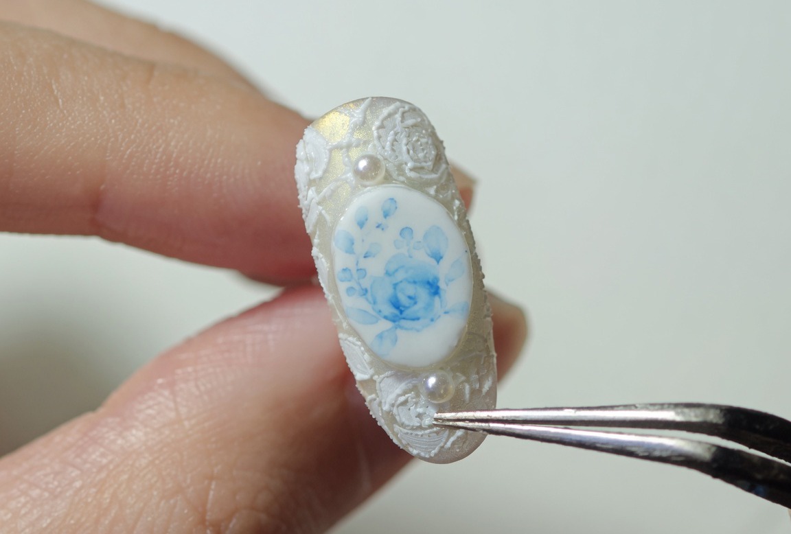 Gros plan dun ongle décoré de roses bleues et de dentelle blanche, tenu par une pince, prêt pour la catalysation finale.