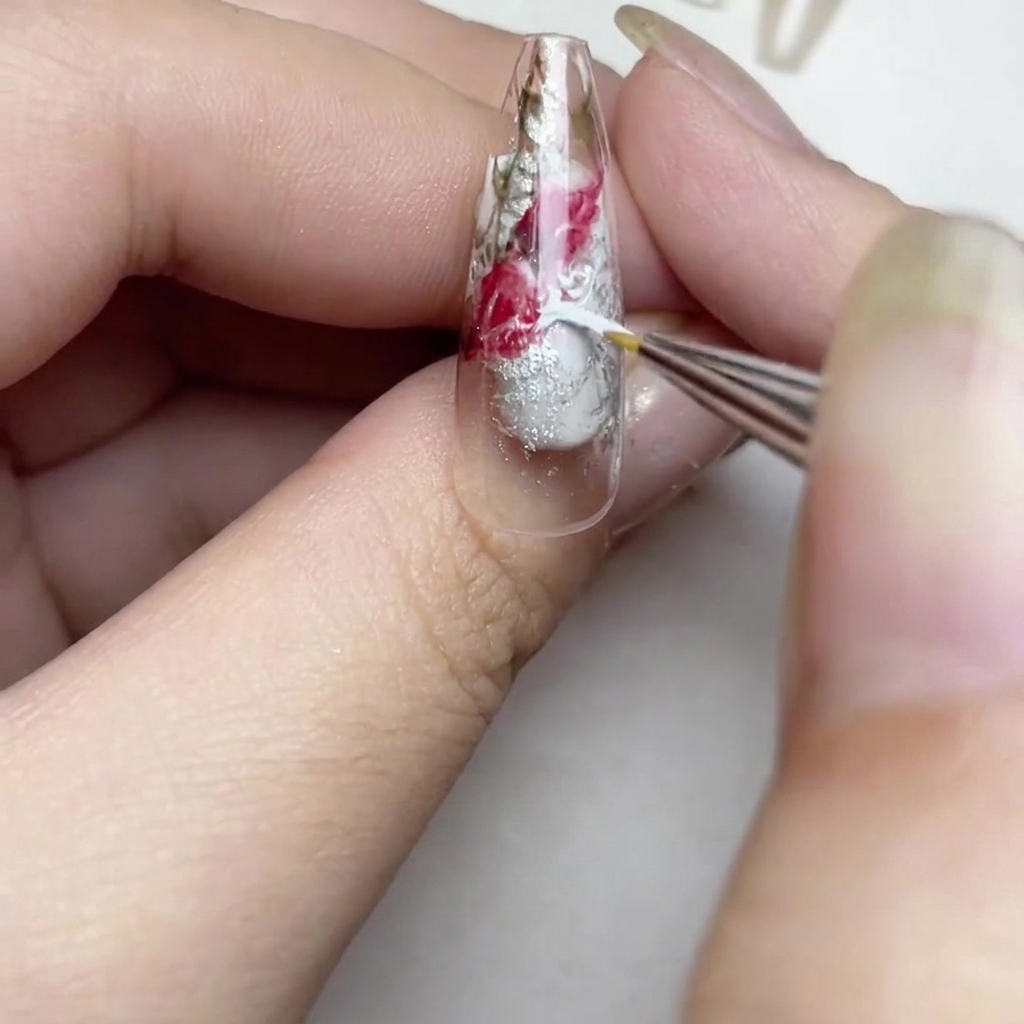 Gros plan sur un ongle montrant un pinceau ajoutant des traits de peinture blanche pour créer un effet de rose aquarelle douce dans les espaces transparents.