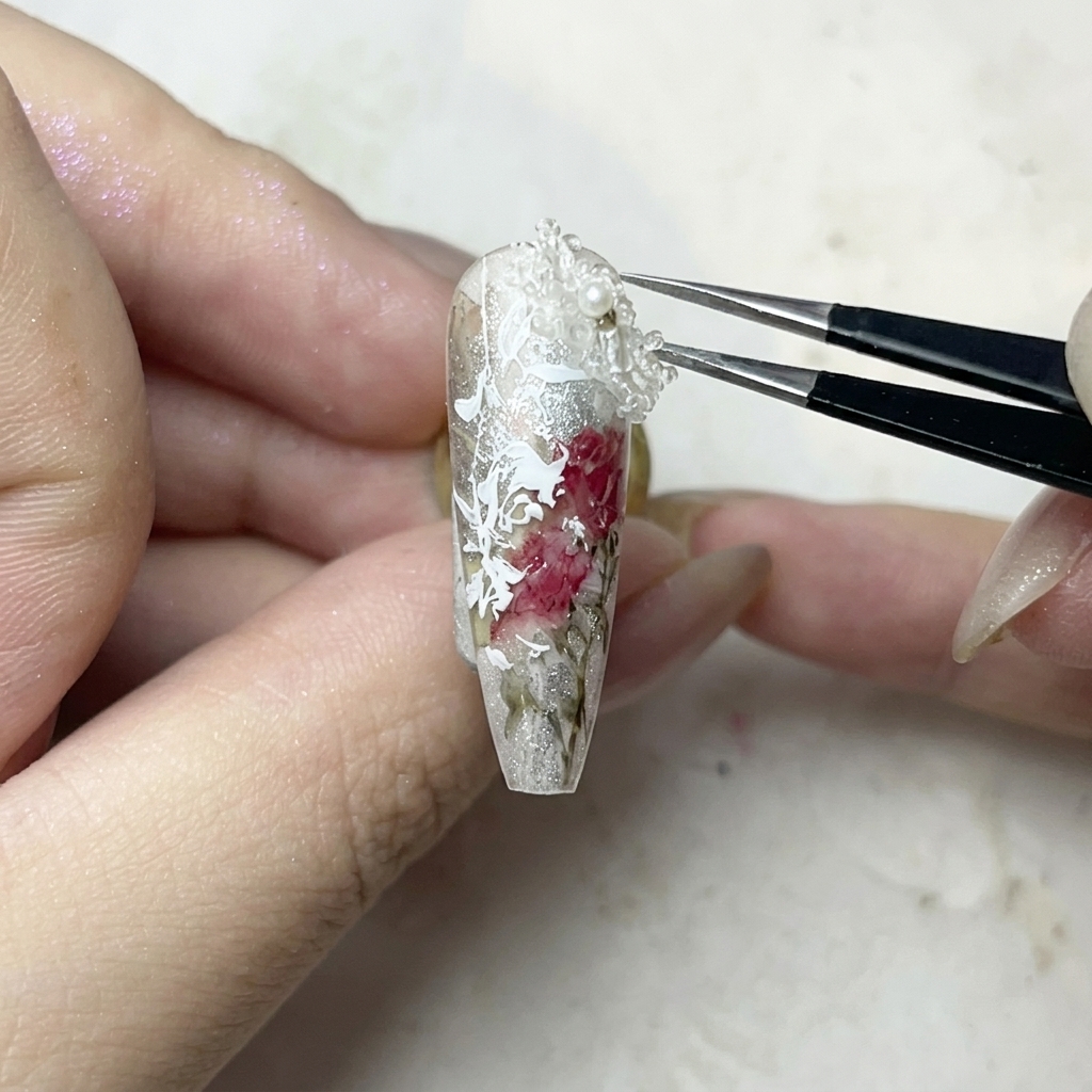 Une pince déposant soigneusement un petit charm floral 3D blanc sur un point de colle gel transparente sur un ongle.