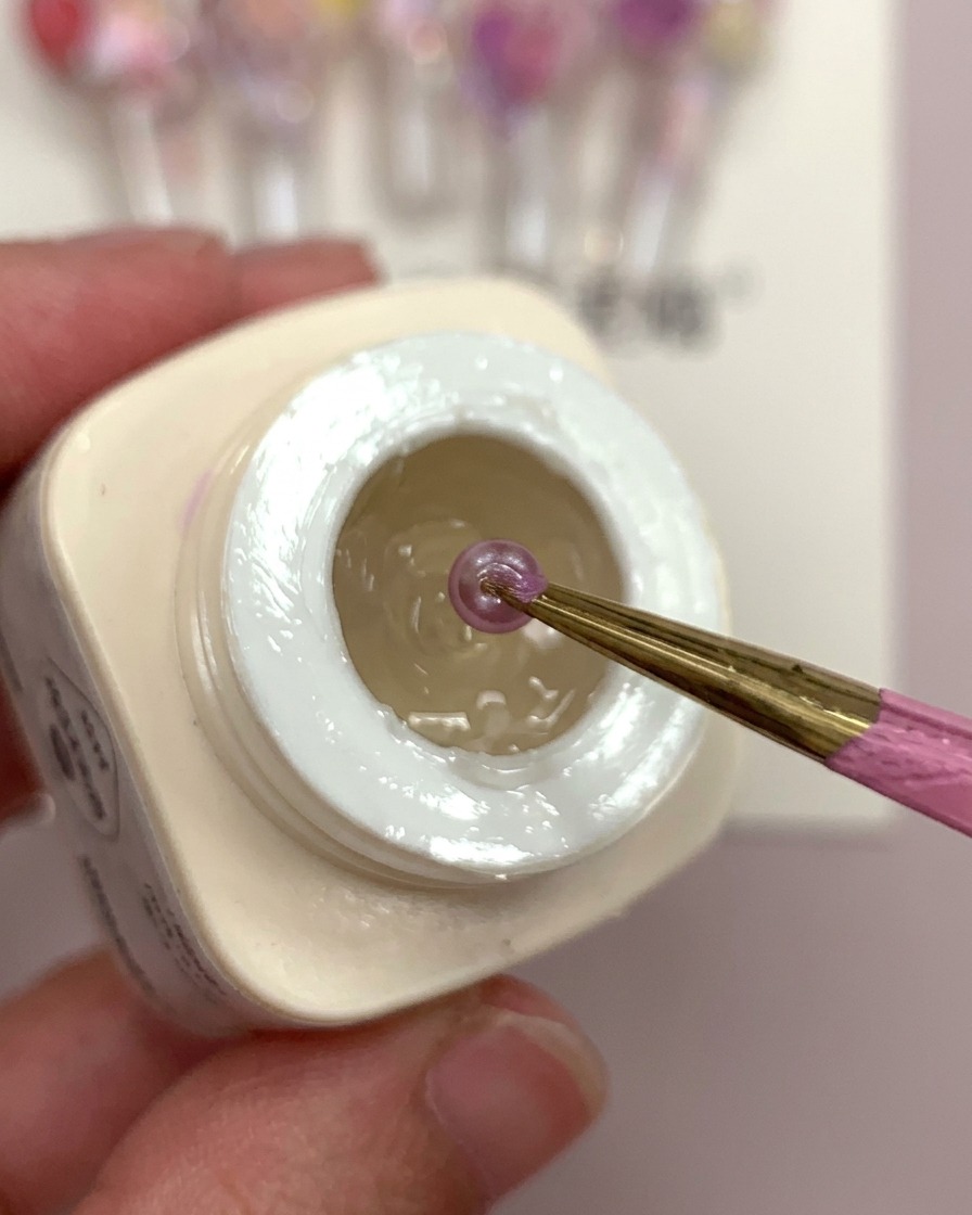 Une main tenant un tip avec une goutte de colle givrée et des boules de fleurs violettes en cours de placement.