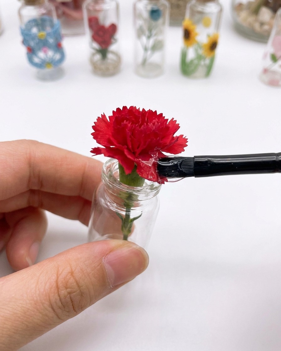 Un petit pinceau appliquant un top coat transparent sur la surface dune fleur dœillet rouge fixée sur un bijou dongle.