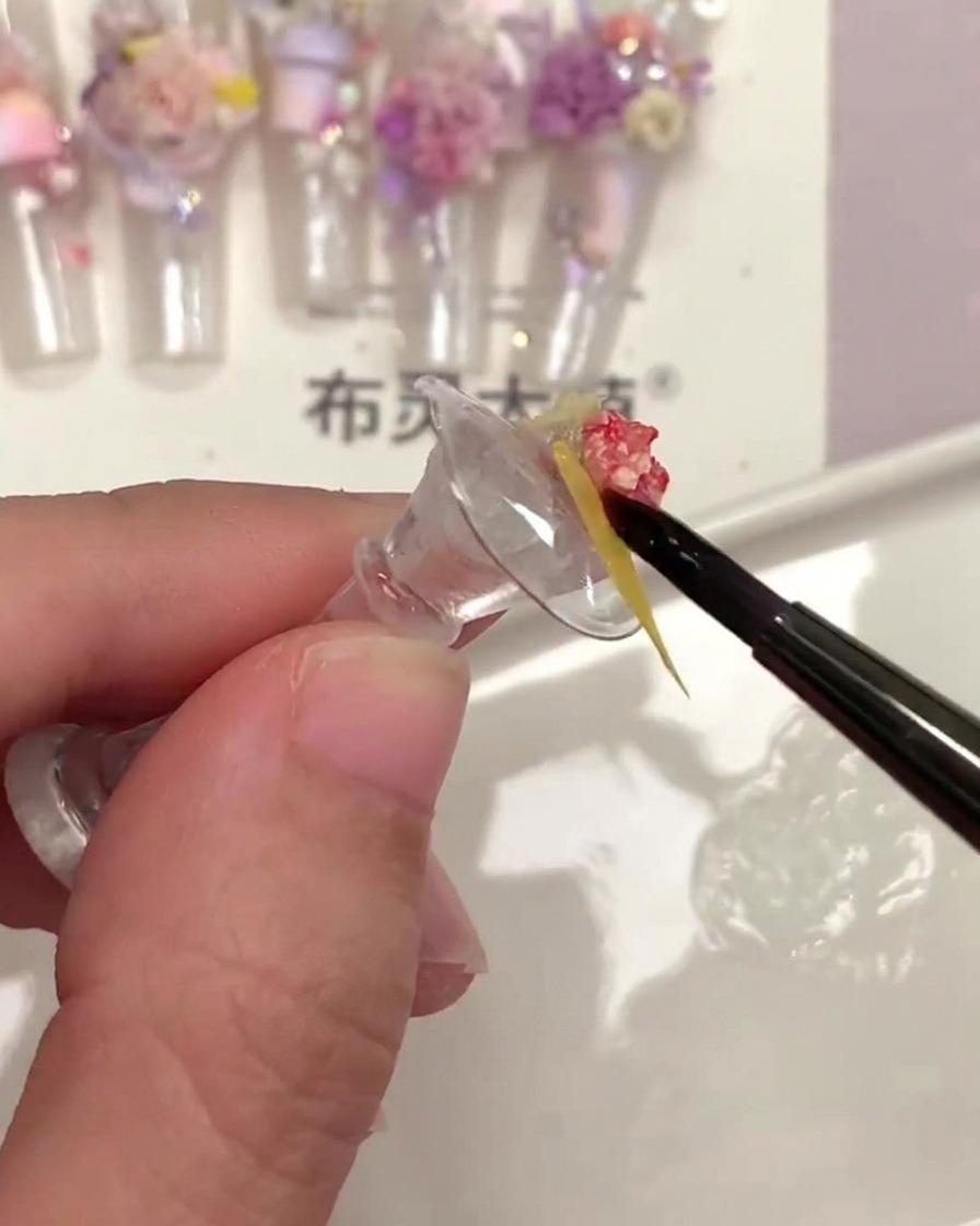 Un pinceau à ongles fin appliquant du top coat transparent sous une petite pièce de fleur séchée jaune.