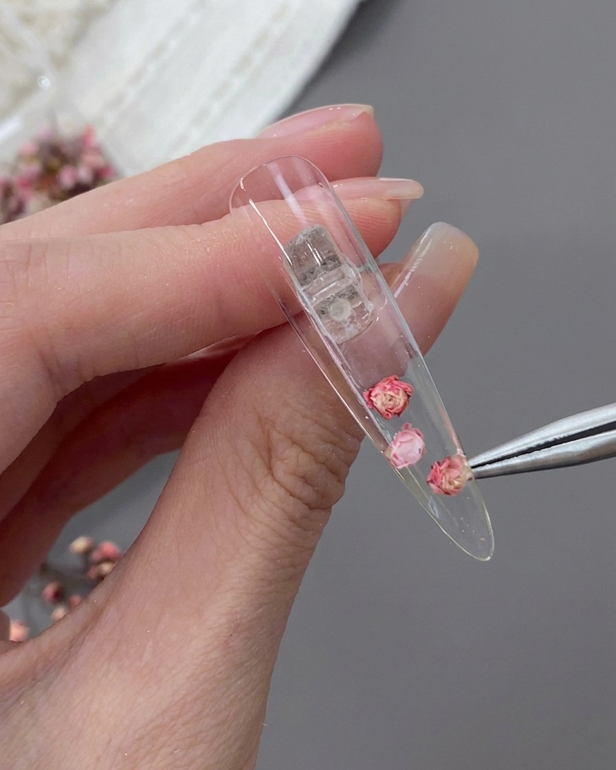 Une pince de précision plaçant une petite fleur séchée rose à côté de la rose centrale à lintérieur dune capsule stiletto.
