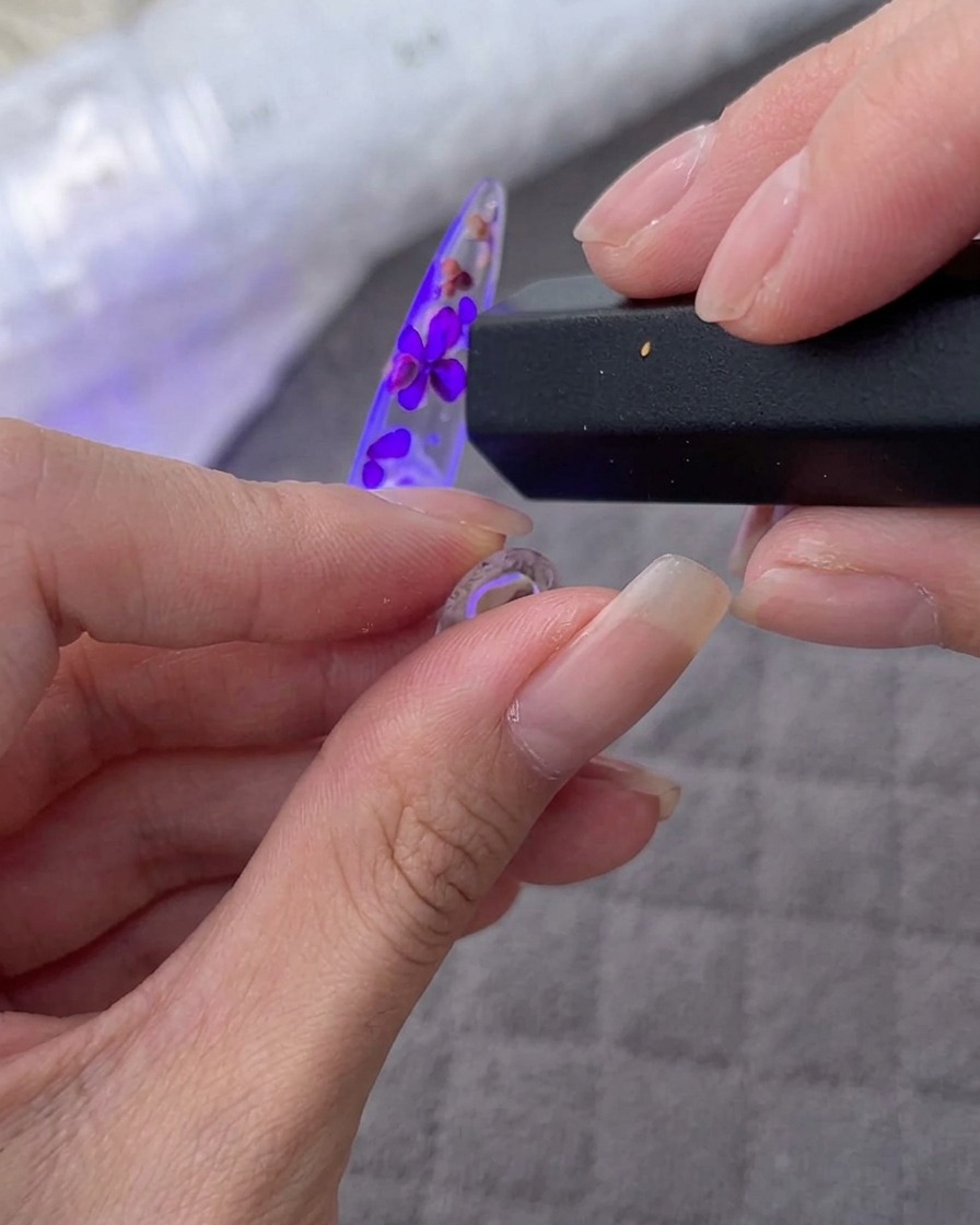 Une main tenant une lampe UV/LED portable au-dessus dune capsule décorée de fleurs et de strass.