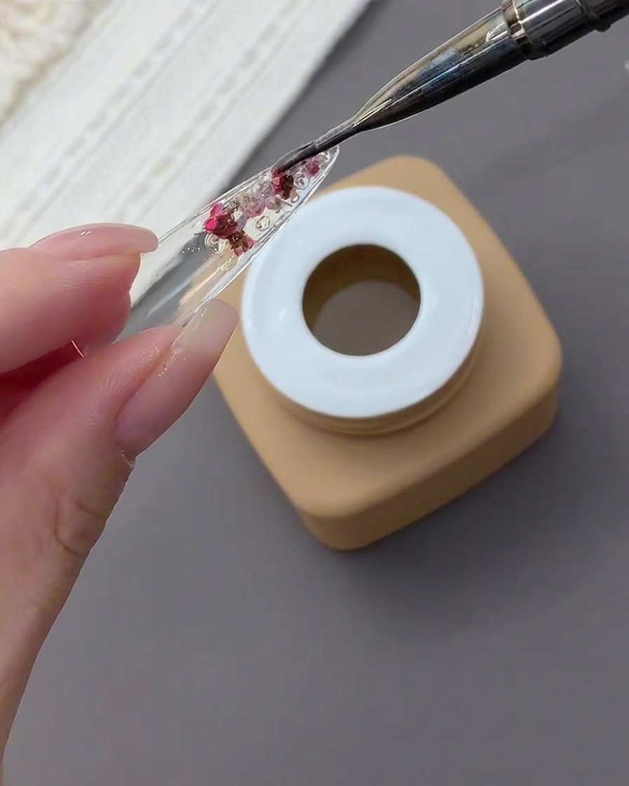 Un pinceau nail art appliquant une épaisse couche de gel transparent sur les fleurs et strass encapsulés au dos de la capsule.
