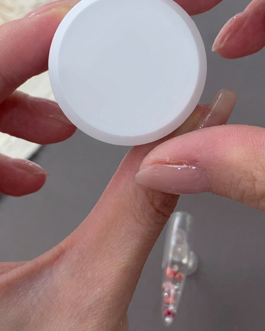 Une main tenant une éponge et tapotant du gel blanc laiteux sur le bord arrière dune capsule transparente.