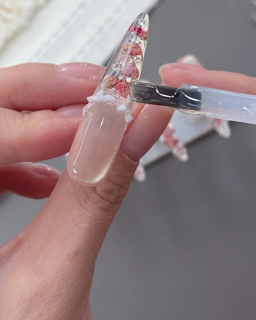 Un ongle décoré avec un nœud blanc, des perles et des strass en cours de finition.