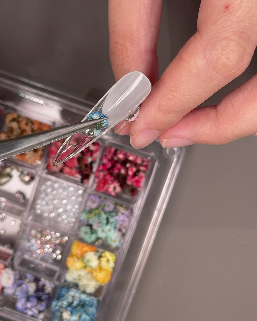 Une pince déposant une petite fleur séchée bleue sur le dessous de la capsule.
