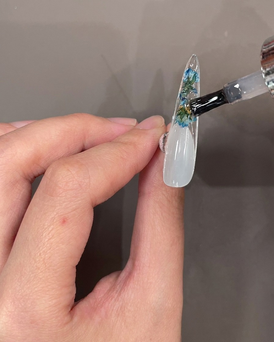 Un pinceau à ongles appliquant une couche de top coat transparent sur les fleurs et strass sous longle.