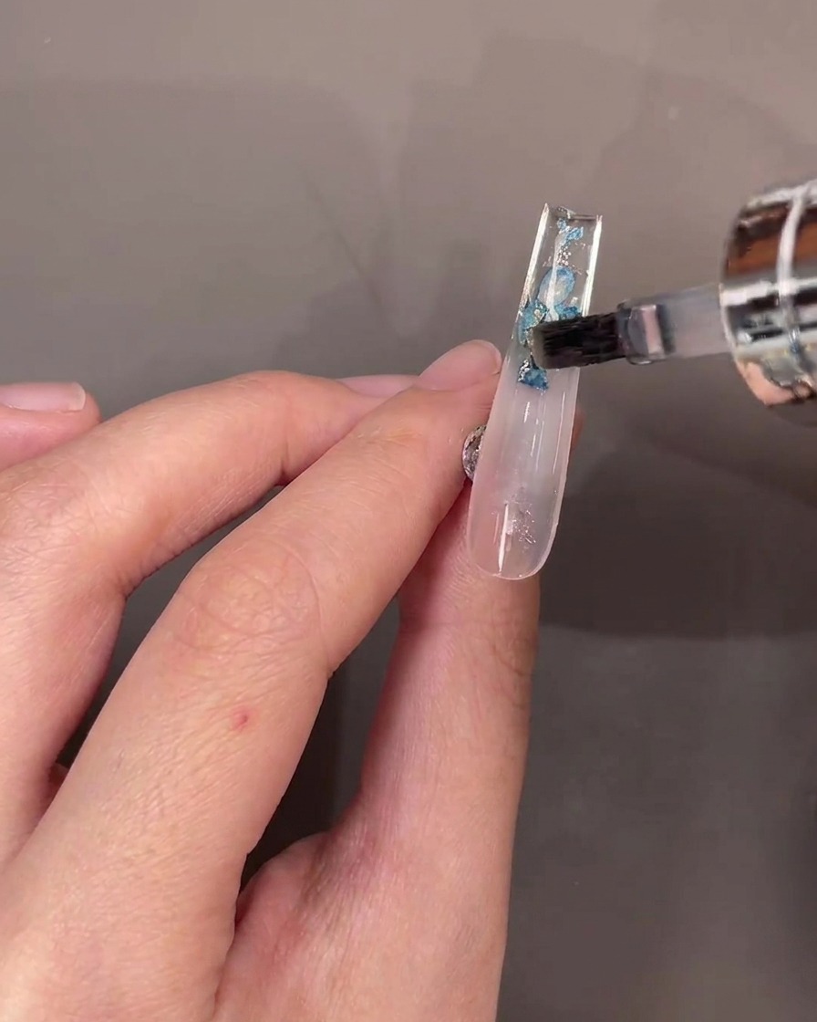 Un pinceau appliquant une couche de top coat transparent sur les fleurs sous longle.