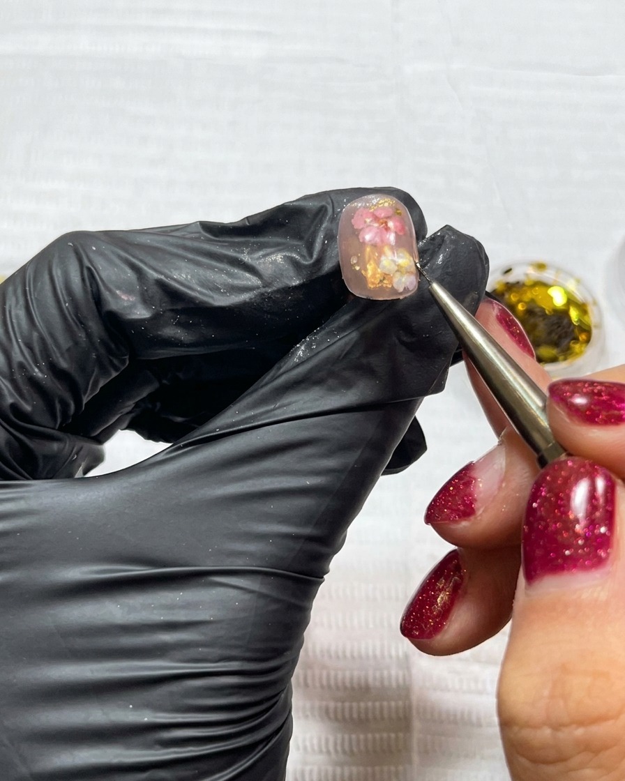 Un pinceau appliquant des flocons de feuilles dor sur un ongle décoré de fleurs.