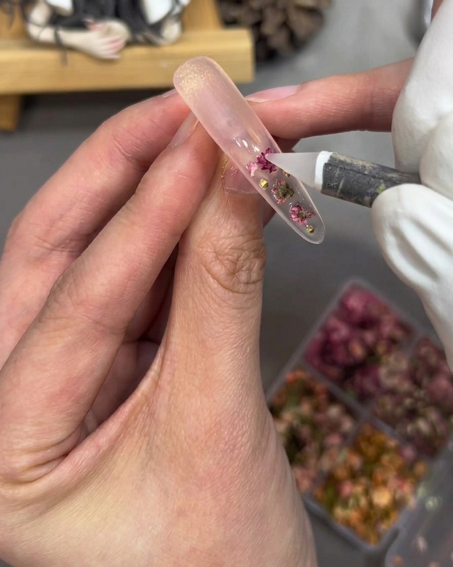 Gros plan sur lutilisation dun outil pour placer de petites perles dorées et des strass roses sur un ongle.