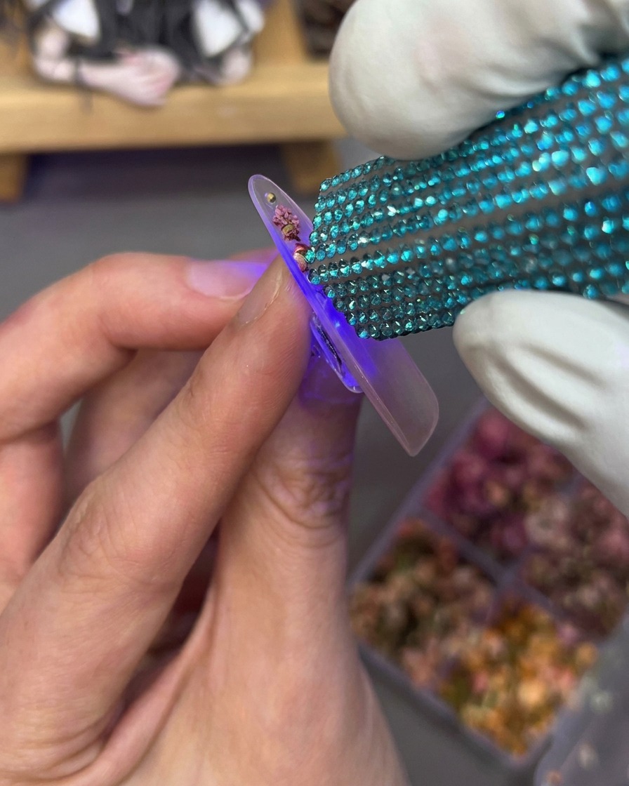 Une main tenant une mini lampe UV au-dessus dun ongle décoré de fleurs et de strass.