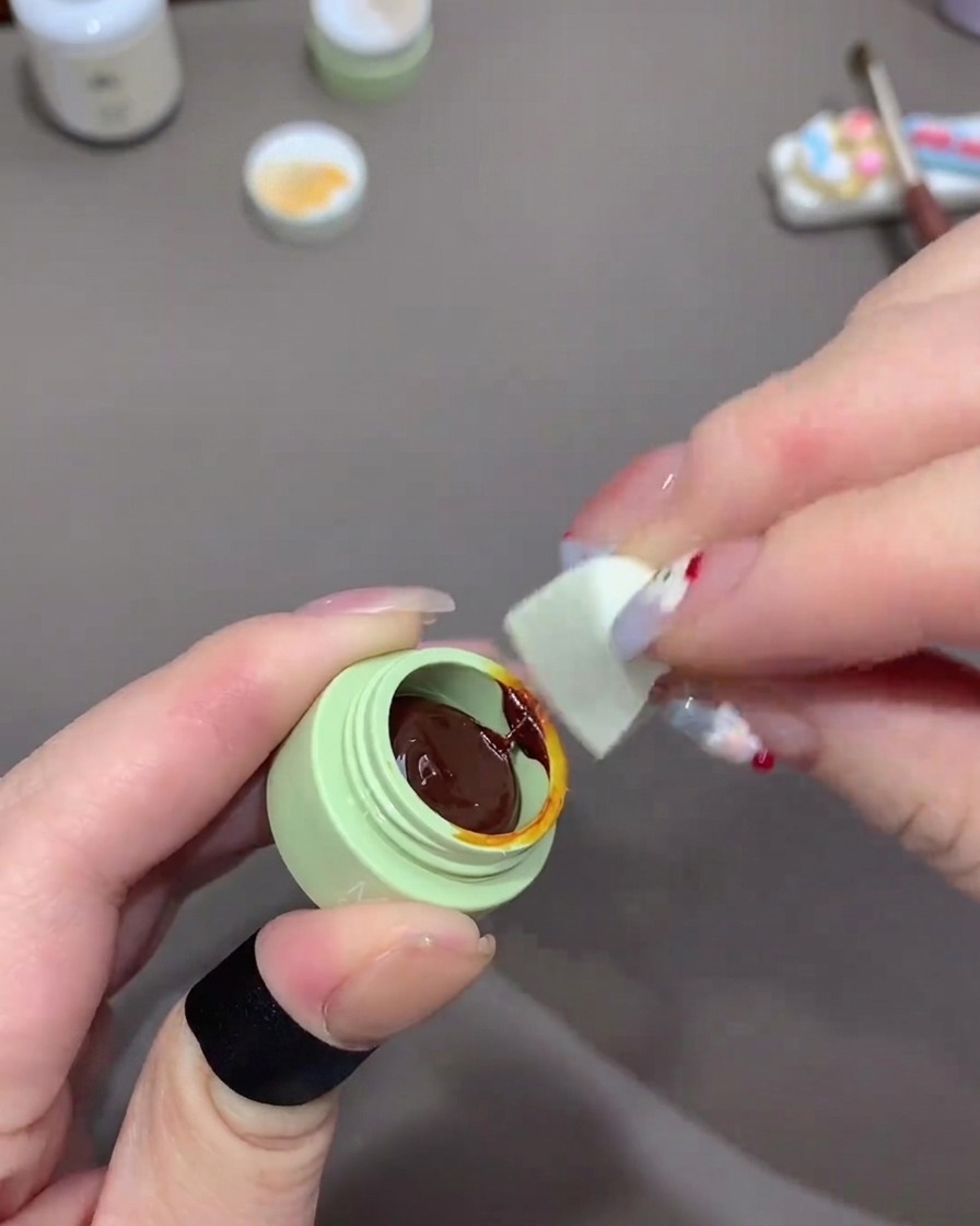 Une main utilisant un morceau déponge à maquillage blanche pour prélever du vernis gel orange foncé dans un pot.